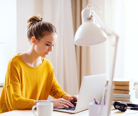 Junge Frau in gelbem Pullover sitzt an einem Tisch und arbeitet am Laptop